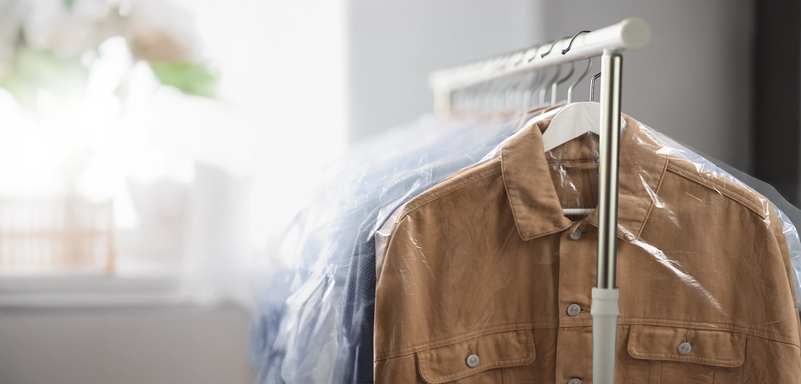 A tan shirt hangs on a rack with other clothes covered in plastic, suggesting they have been dry cleaned.