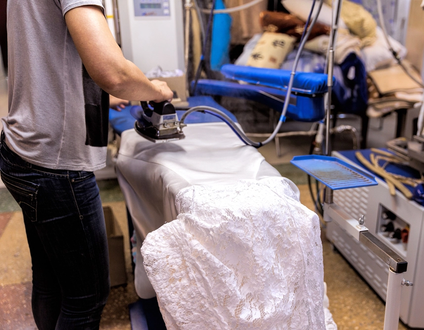 A person irons a white lace fabric on an ironing board in a room with various equipment and textiles.