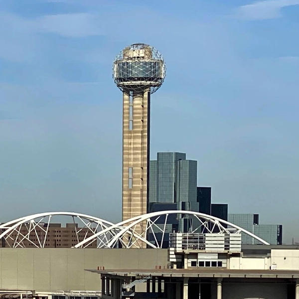 A tall tower with a geodesic dome at the top stands in front of modern office buildings and an arched white bridge under a blue sky.