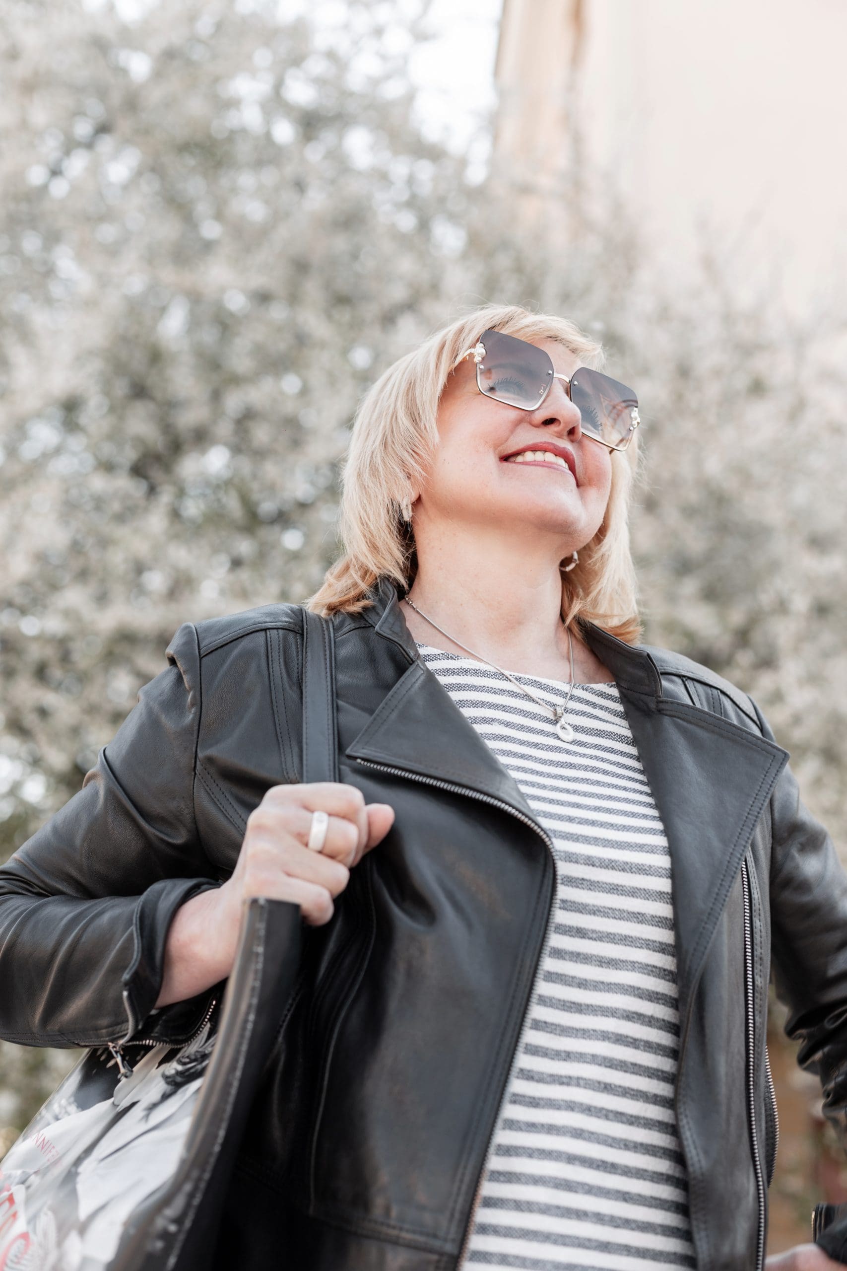 Woman wearing sunglasses and a black leather jacket stands outdoors, smiling and holding a bag, with blurred trees in the background.