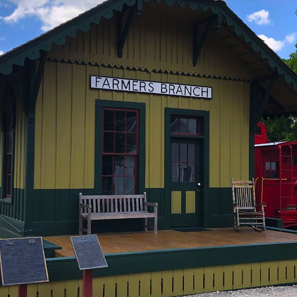 A historic train depot building labeled "Farmers Branch" with benches and informational plaques in front.