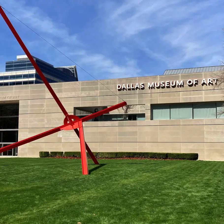 Exterior of the Dallas Museum of Art with a large red metal sculpture on a green lawn under a blue sky.