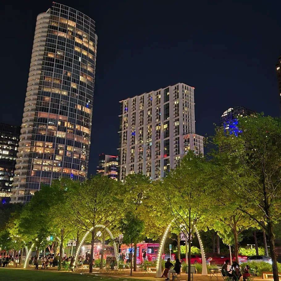 City park at night with people sitting on benches, illuminated arches, tall buildings, and trees in the foreground.