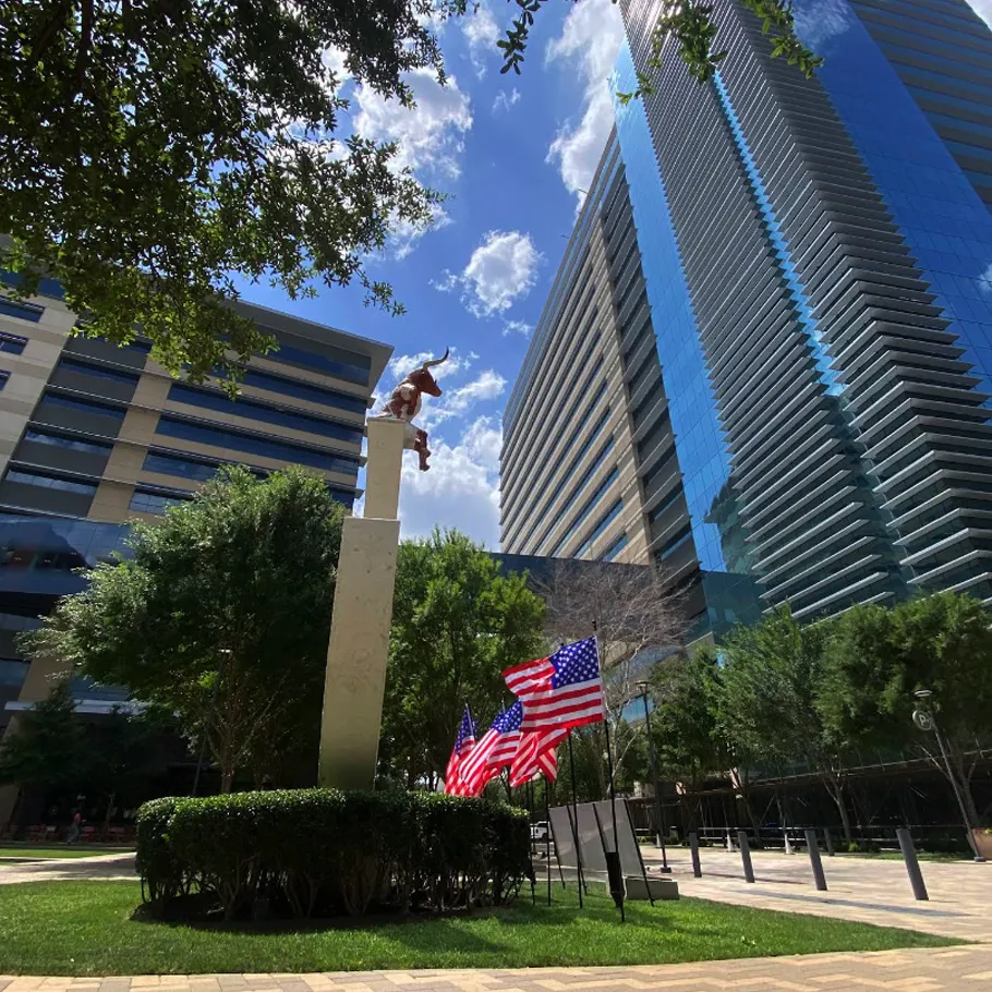 A tall statue on a pedestal stands near several American flags, surrounded by trees and modern office buildings under a partly cloudy sky.