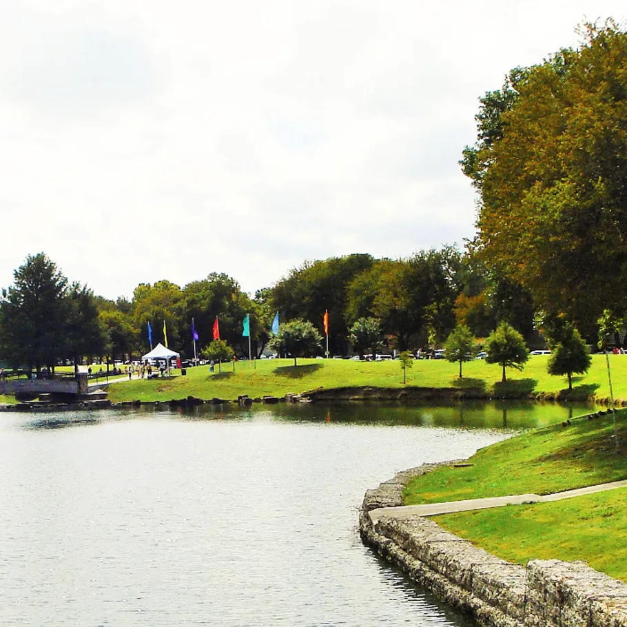 A park with a grassy shoreline, trees, and colorful flags near a calm body of water. A few tents and people are visible in the background.