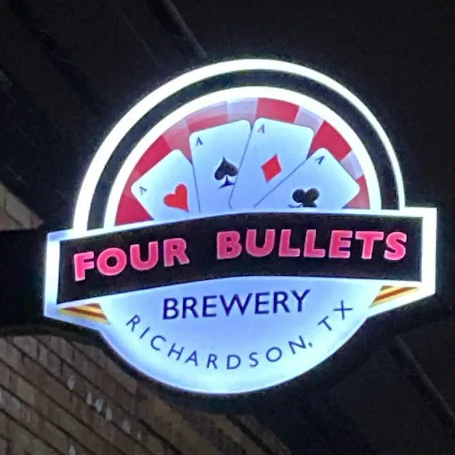 A round illuminated sign for Four Bullets Brewery in Richardson, TX, featuring four playing cards: ace of spades, ace of hearts, ace of diamonds, and ace of clubs.