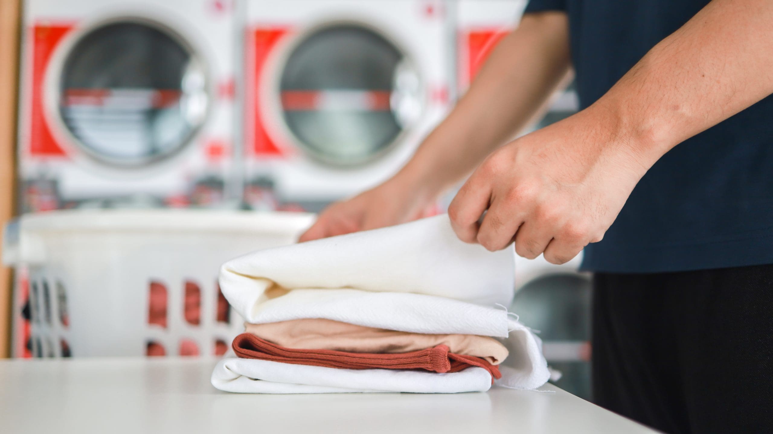 A person is folding clean laundry on a table in a laundromat, with washers or dryers and a laundry basket visible in the background.