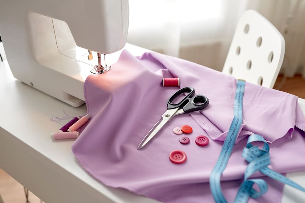 A sewing machine with lavender fabric, scissors, pink buttons, spools of thread, and a blue measuring tape on a white table and chair.