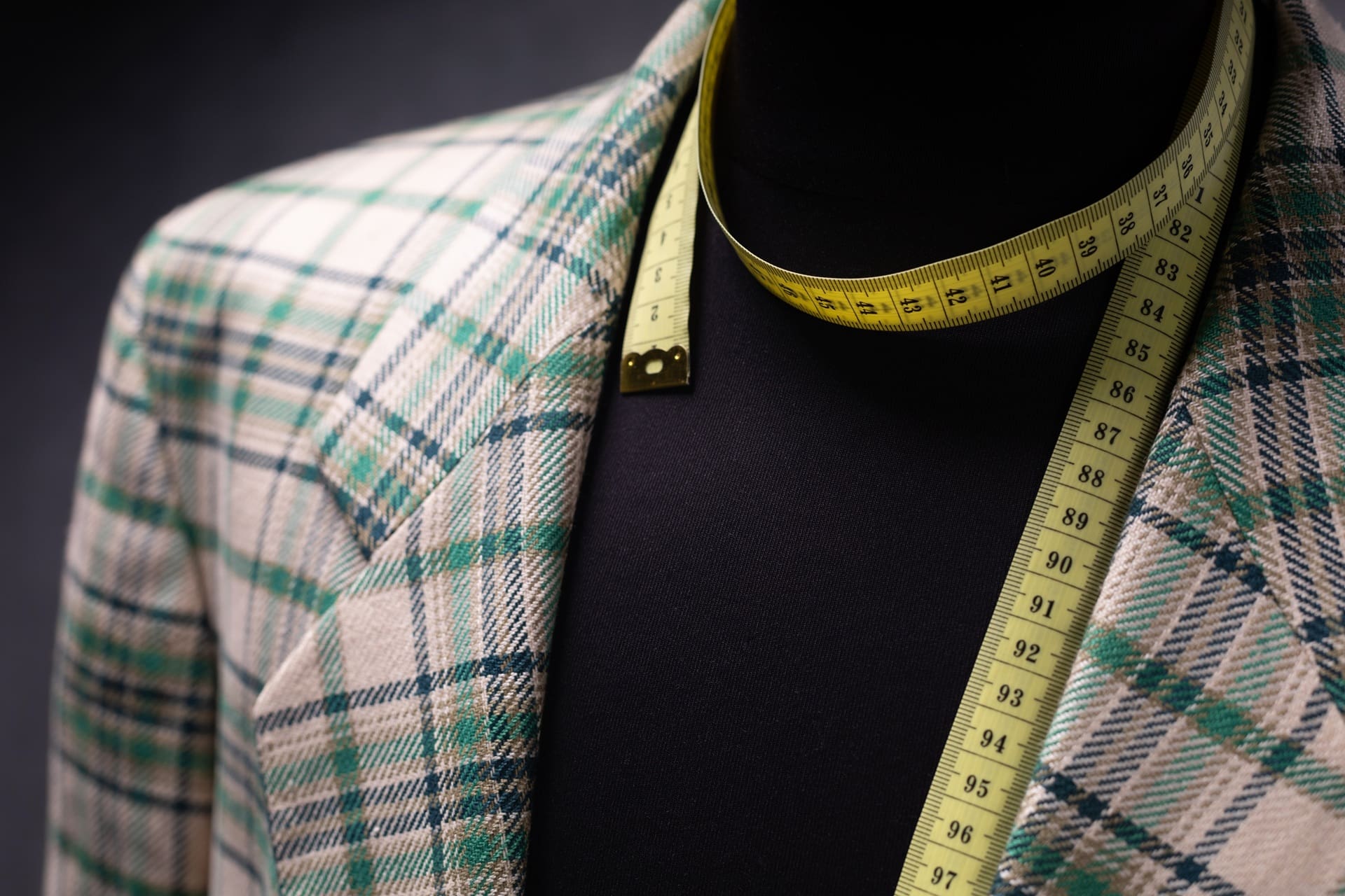 A plaid blazer on a black mannequin with a yellow measuring tape draped around the neck.