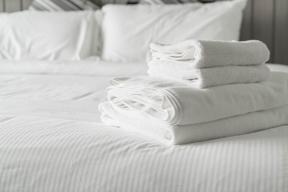 A neatly folded stack of white towels is placed on a made bed with white linens and pillows in the background.
