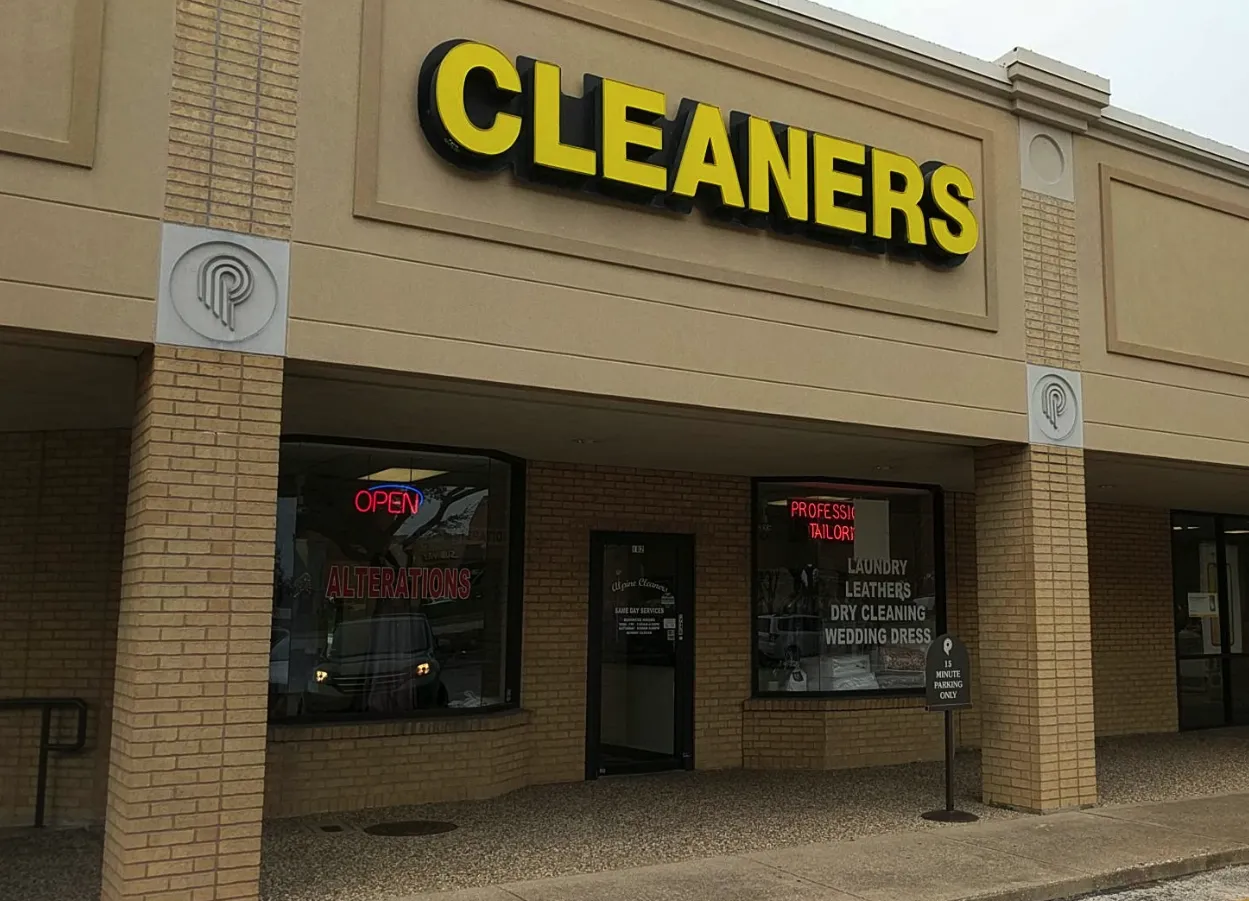 Storefront of a dry cleaners with a large yellow "CLEANERS" sign, neon "OPEN" and "ALTERATIONS" signs, and a list of services on the window.