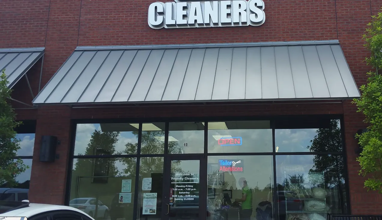 Storefront of a dry cleaning business with a "CLEANERS" sign, glass windows, a visible "OPEN" sign, and people inside. Trees and a car are partially visible outside.