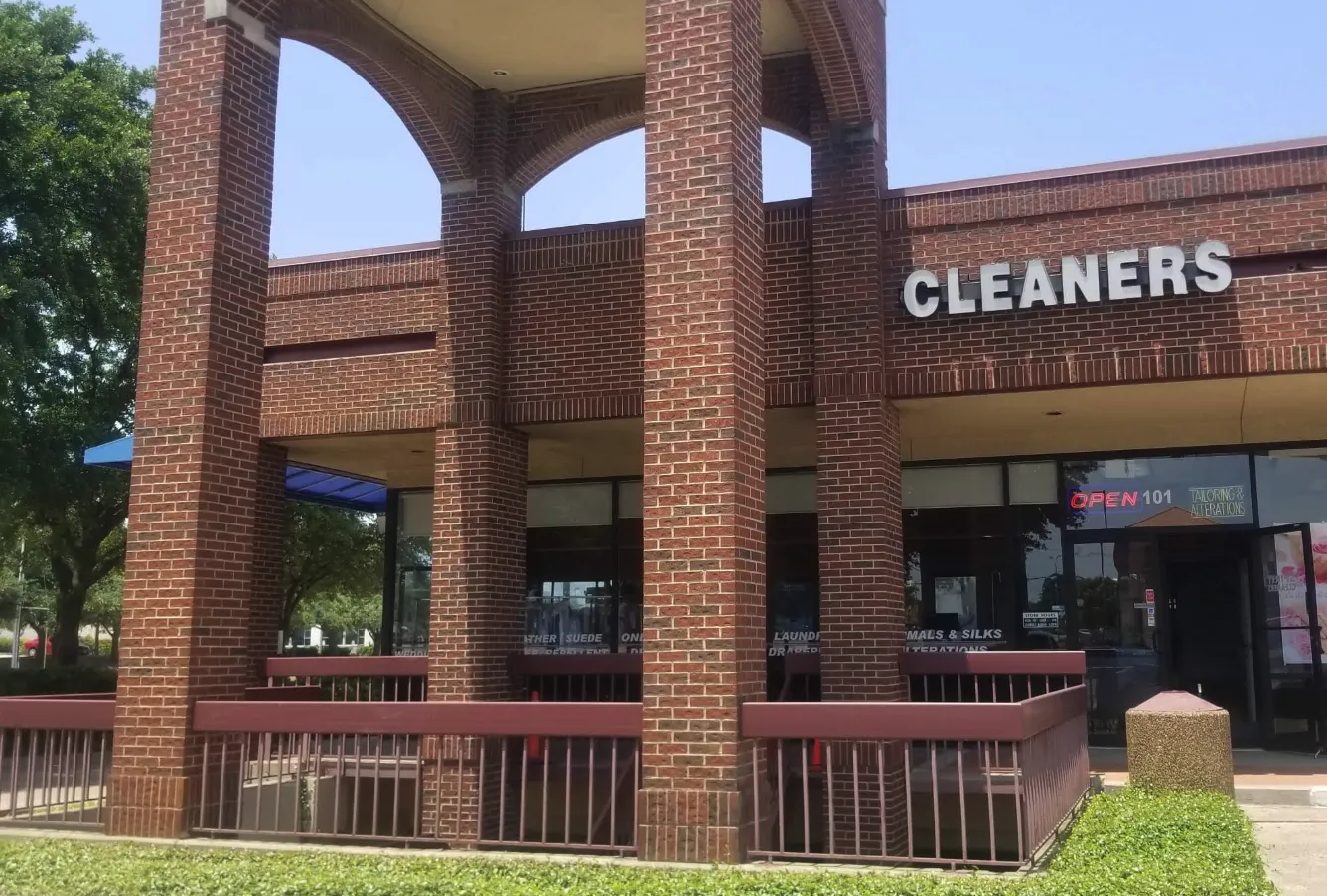 A brick commercial building with columns, partial signage reading "CLEANERS," and an "OPEN" sign in the window. There is an empty, fenced-in patio area in front.