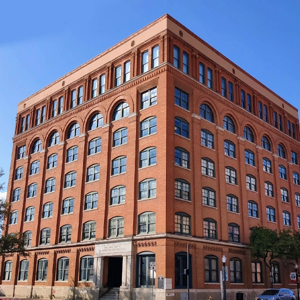 A large, historic red brick building with arched windows on each floor, under a clear blue sky.