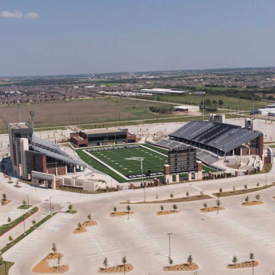 Aerial view of a large, modern football stadium with empty stands and parking lot, surrounded by open fields and residential areas in the distance.