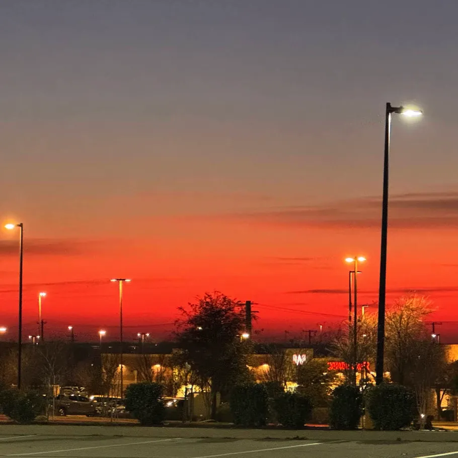 Parking lot at sunset with orange and red sky, streetlights turned on, and buildings with illuminated signs in the background.