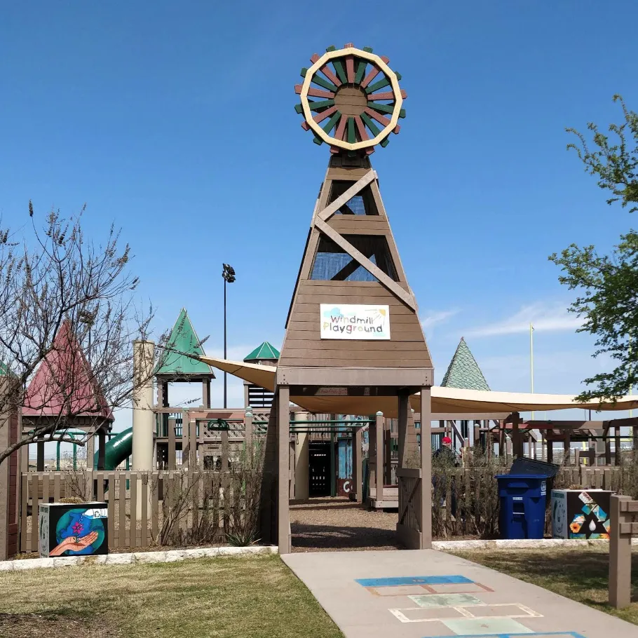 Wooden windmill structure marks the entrance to a playground with slides and climbing areas under a clear blue sky.