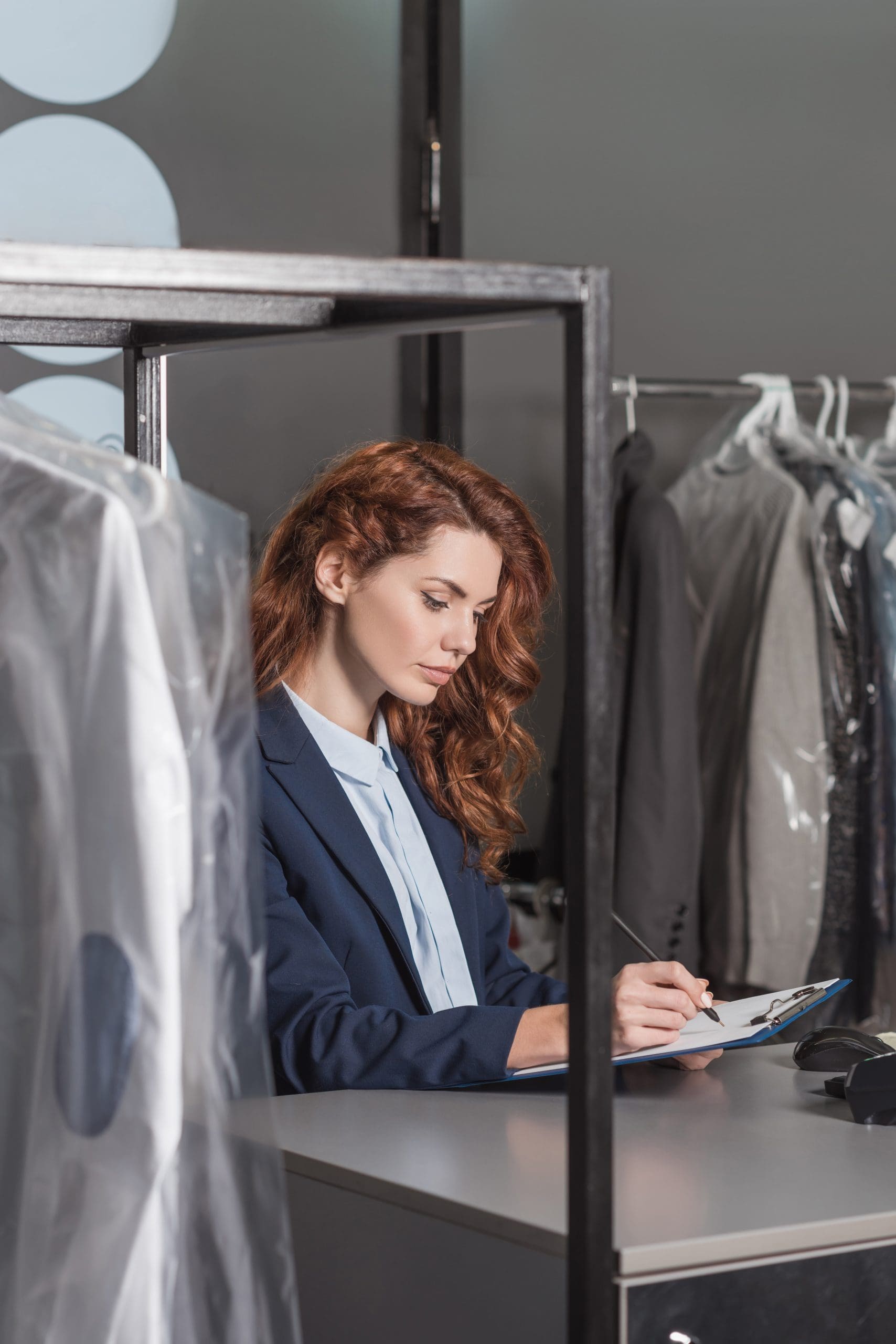 A woman in a suit sits at a counter, writing on a clipboard, with hanging clothes and garments covered in plastic in the background.
