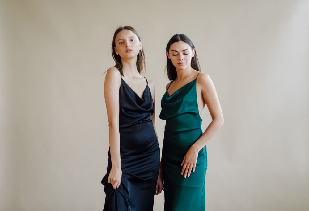 Two women stand side by side against a plain beige background, wearing sleeveless satin dresses—one in black and the other in dark green.