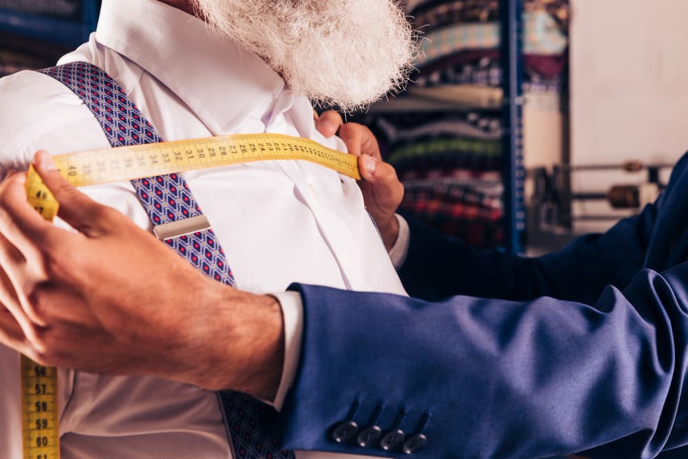 A tailor in a blue suit measures the chest of a man wearing a white shirt and suspenders with a yellow measuring tape in a fabric shop.