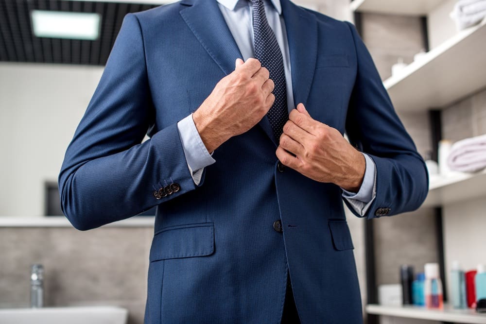 A man in a blue suit adjusts his jacket in a bathroom, with shelves and toiletries visible in the background.