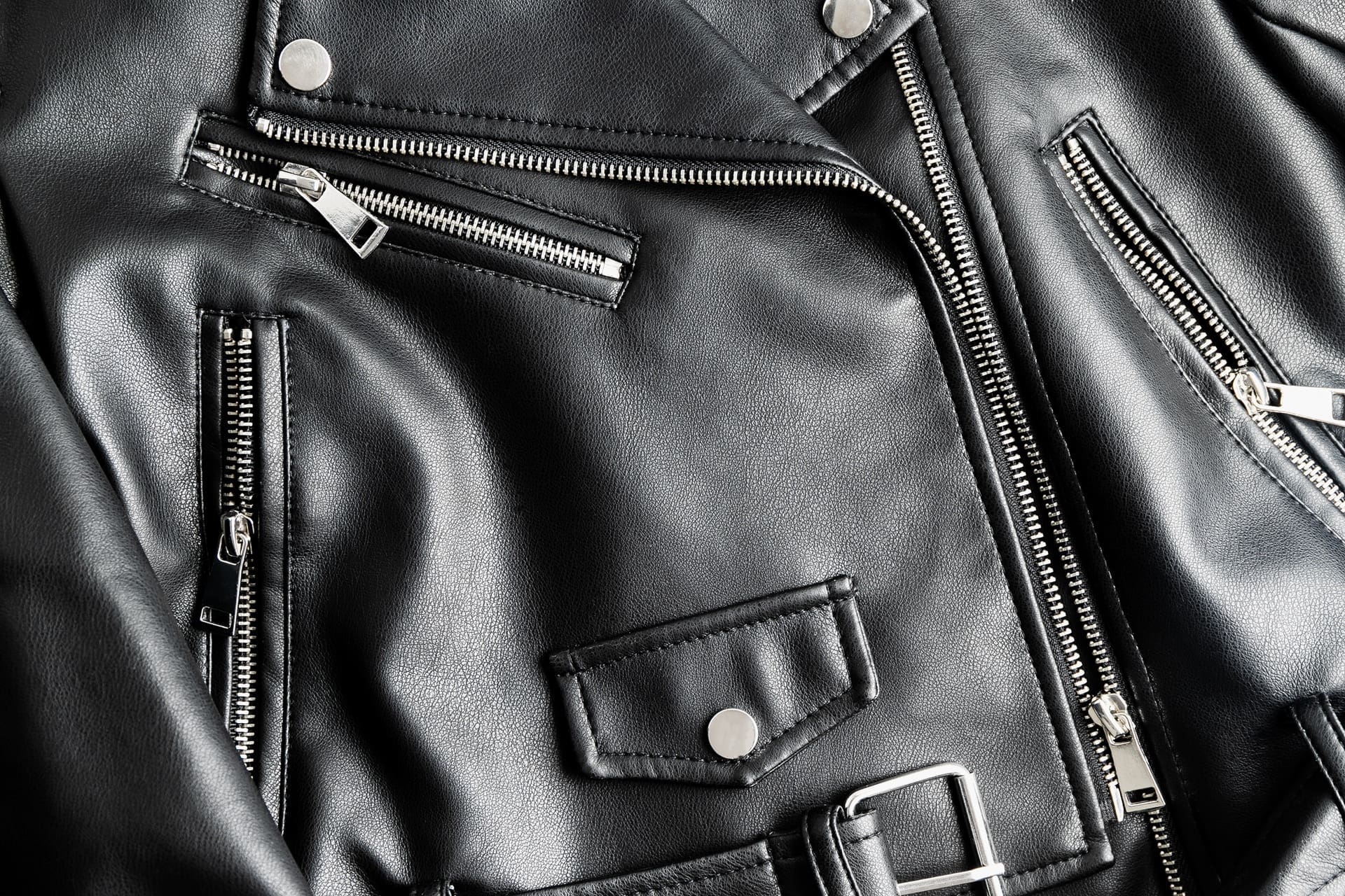 Close-up of a black leather jacket featuring multiple silver zippers, snap buttons, and stitched details.