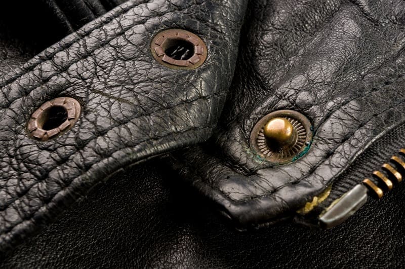 Close-up of a worn black leather jacket showing textured material, metal snap buttons, and part of a brass zipper.