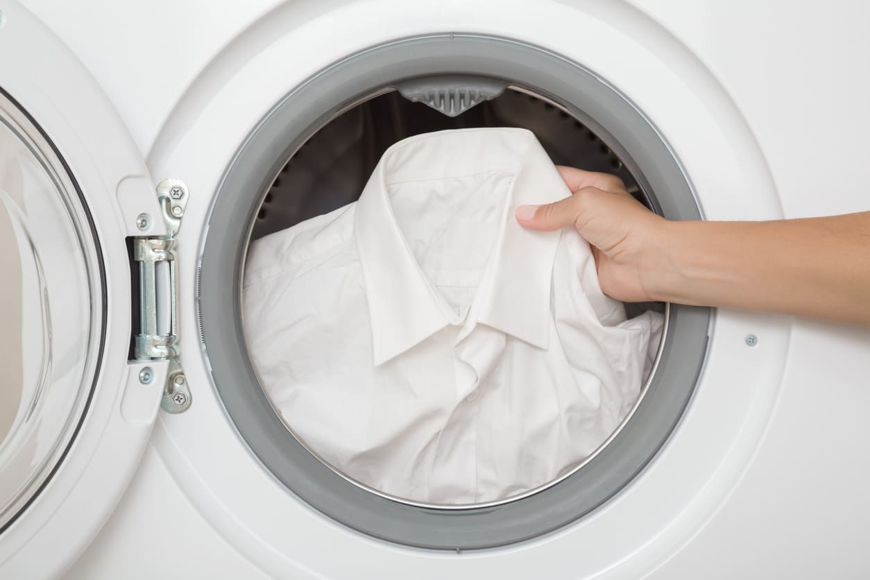A hand is placing a white dress shirt into the drum of a front-loading washing machine with the door open.